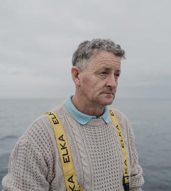 A photograph of a fisherman in a white aran jumper on a boat with a dejected expression