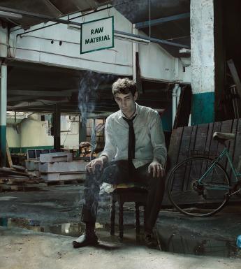 A painting of a young man, sitting on a chair in a dark abandoned warehouse, surrounded by rubbish.
