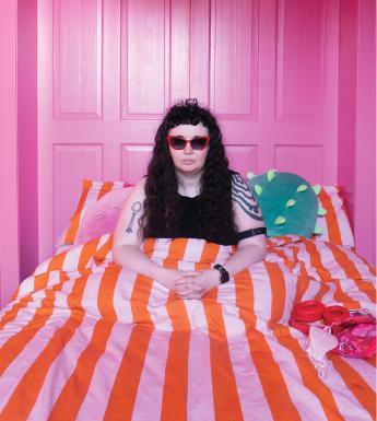A young girl with long black hair, sits up in a bed with a pink and orange striped duvet, in a pink room.