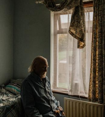 Photograph of an older man with a white beard, sitting in a small room in blue pyjamas