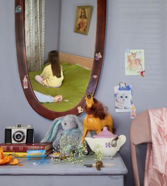 Photograph of a young girl with her back to us reflected in a mirror in front of an outstretched arm holding rosary beads. 