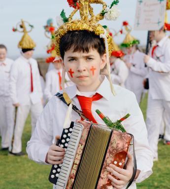 A young boy wears a traditional 'Biddy Boy' outfit and holds an accordion.
