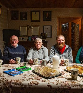 A photograph of three men and two women, sitting behind a table covered in cups of tea and sandwiches.