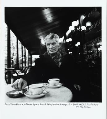 Black and white image of an older male figure sitting at a Parisian cafe, with two coffee cups on the table in front of him.