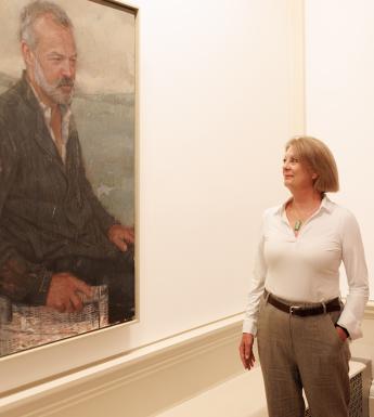 A woman stands with one hand in her pocket looking at a portrait hanging beside her, of a man seated. 
