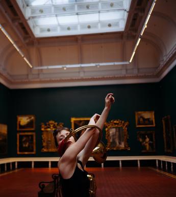 A figure stands in a gallery filled with paintings, arms outstretched through two metal rings.