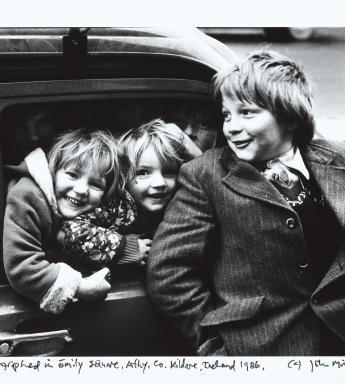 Black and white photograph of three young children laughing in front of a car.