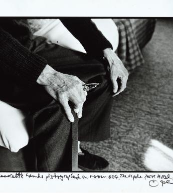 Black and white photograph focusing on an older man's hands as he sits on the edge of a bed, resting his hands on his knees