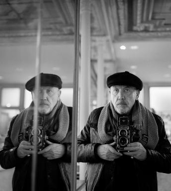 Black and white self-portrait photograph of a man reflected in a mirror, wearing a scarf and black hat and holding a Rolleiflex camera