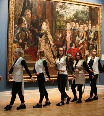A group of nine women wearing cream jackets walk in sequence through a Gallery, in front of a large painting
