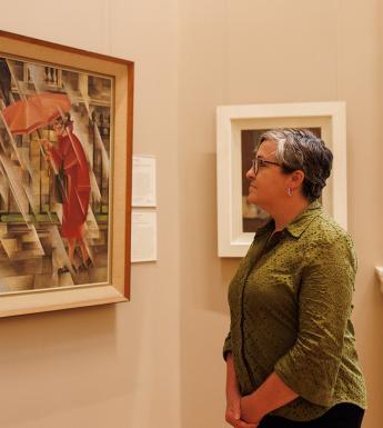 A woman in a green shirt stands in a gallery looking at a painting on the wall of a woman in red with an umbrella over her head