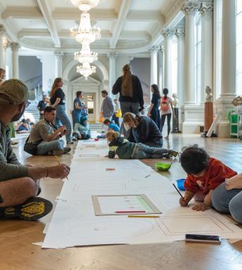 A group of adults and children sit on the floor of a large hall, drawing on a long sheet of paper running the length of the room.