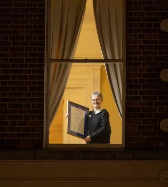 A figure stands at a window, holding a framed certificate.
