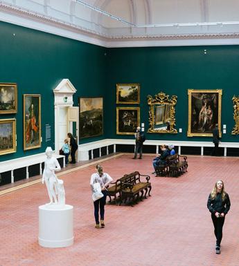 Photo of people walking through a green gallery room with gilt-framed paintings covering the walls and a white marble statue standing in the middle of the room.
