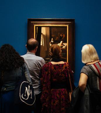 A photograph of people, with their backs to the camera, looking at three gilt-framed paintings on a blue wall.