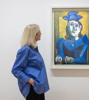 A woman in a blue shirt looks towards Picasso's painting Bust of a Woman with a Blue Hat, hanging on a white gallery wall.