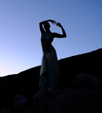 A figure in silhouette stands in a balletic pose, a blue sky and shadowed hills behind them.