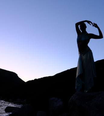 A dancer stands on the banks of a river with her arms linked above her head, silhouetted against a blue sunrise sky. 