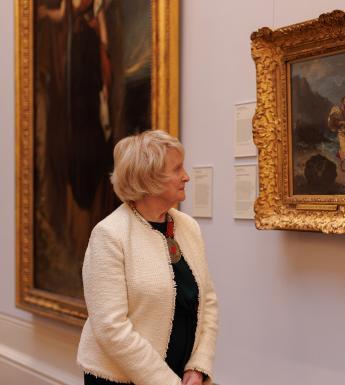 A woman in a white jacket and black dress stands in a gallery looking towards one of the paintings on the wall. 