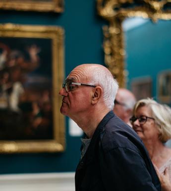 Photo of a man and woman in a gallery room with gilt-framed paintings in the background.