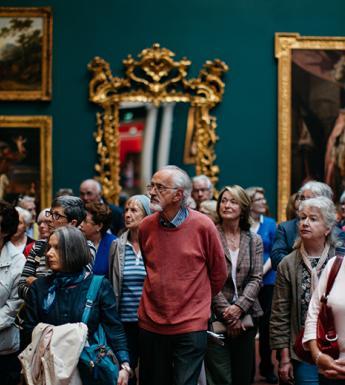 Photo of a group of people on a guided tour in a gallery, with gilt-framed pictures and mirrors on a wall behind them.