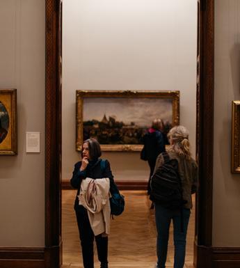 Photo of two women passing each other in a doorway in a gallery room with gilt-framed paintings on the walls.