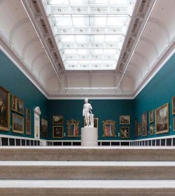 Large green room with high ceilings, a glass ceiling, walls hung with paintings and a white marble statue in the middle of the room.