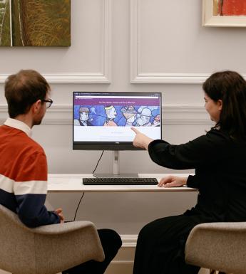 A man and a woman sit in front of a computer. The woman is pointing at an image on the screen.