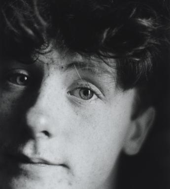 A black and white photograph of a young man's head. His face and curly hair fill most of the frame, and one side of his face is slightly out of frames looking directly at the camera and 