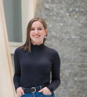 Sarah stands in the Gallery Courtyard. She is smiling, with her hands in her pockets.