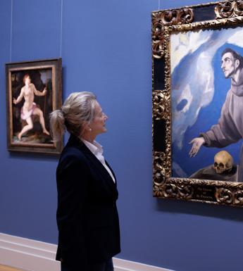 A woman in a black jacket stands gazing at a painting of Saint Francis on a blue Gallery wall