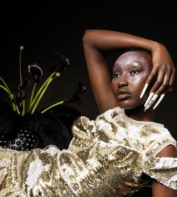 Photograph of woman with nail art, reclining beside plant, against black background.