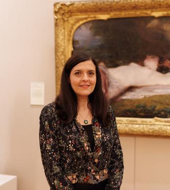 A woman with long dark hair stands in front of a painting in an elaborate gilt frame.
