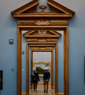 A photograph of a suite of galleries, looking through a series of ornate wooden doorframes. The walls are painted blue, and we can see some paintings hanging. In the distance, some people are walking through the rooms.