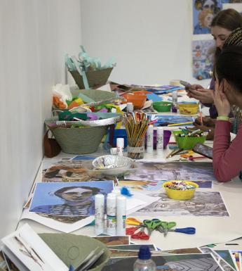 Two people sit at a long table covered in art supplies and materials