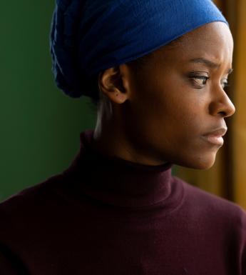 A film still of a young black female figure wearing a burgundy polo neck and a blue headscarf 