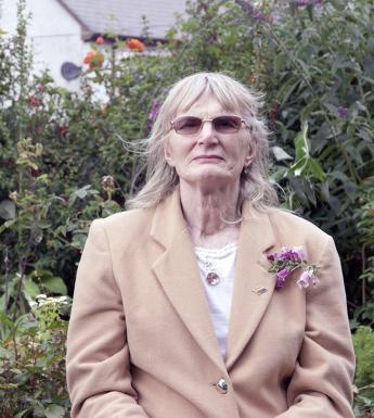 A woman in a beige coloured jacket stands in a garden, wearing dark glasses. On her lapel there is a foxglove; the garden behind is also filled with foxgloves.
