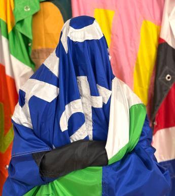 A figure is sitting with their arms folded covered in a large blue and green flag with white writing