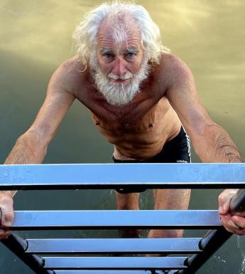 Photograph of an older male figure with long white hair and a beard wearing shorts climbing a ladder out of water