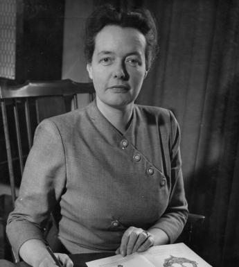 Photograph of Bea Orpen, sitting on a chair, looking at the camera with a book of her artwork open in front of her.