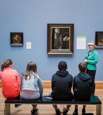 A woman wearing a face shield standing in front of Vermeer's Woman Writing a Letter with her Maid, speaking to a group of 5 school students and their teacher.