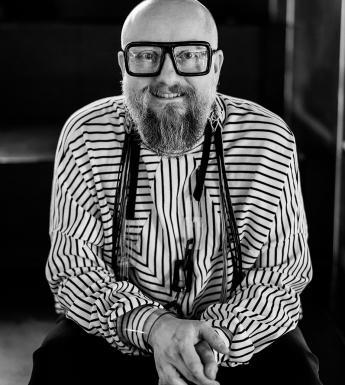 Black and white photo of a man who is bald, wearing thick rimmed black glasses, a long beard and a black and white striped top