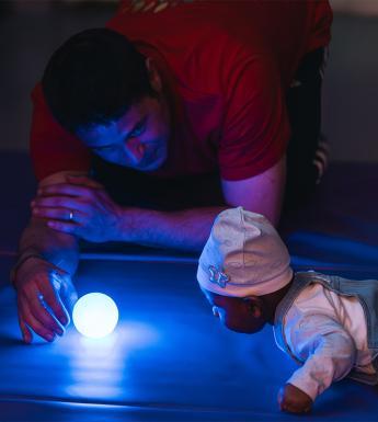 A man holds a bright blue round light on the floor in front of a baby who is crawling towards it.
