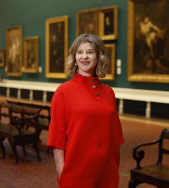 A woman in a red dress stands in a Gallery hung with large oil paintings.