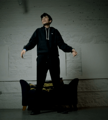 A film still of a person wearing black clothes dancing in front of a black armchair in a room with white walls