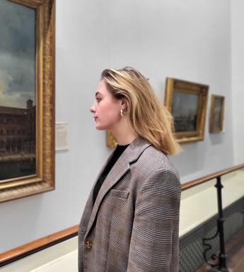 A woman stands in front of a large oil painting, looking closely at it.