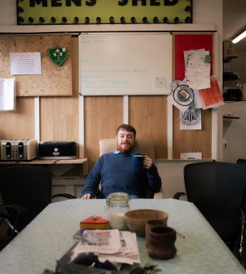 A man in a blue jumper sits at the end of a formica table  under a large yellow sign that says 'Mens Shed'