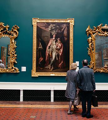 A man and a woman look at art in a grand room filled with painting and antiques.