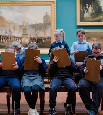 A group of school children in uniform draw on clipboards in a grand gallery filled with paintings.