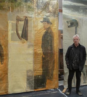 Artist Hughie O'Donoghue standing in his studio in front of two large artworks depicting Michael Collins and Wuffa.